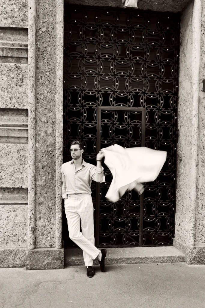 Black-and-white portrait of a man leaning in a doorway with an ornate iron gate, wind-blown white jacket, striped trousers, Milano Fashion Week